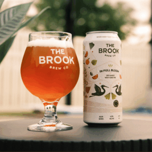 In Full Bloom can and poured glass of beer from The Brook Brew Co. on a patio table with greenery in the background