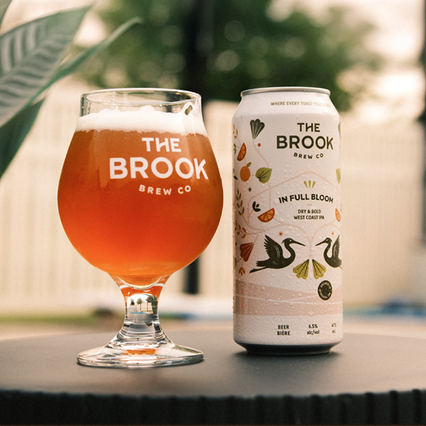 In Full Bloom can and poured glass of beer from The Brook Brew Co. on a patio table with greenery in the background