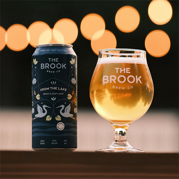 From the Lake Lager can and poured glass of beer from The Brook Brew Co. with warm bokeh lights in the background