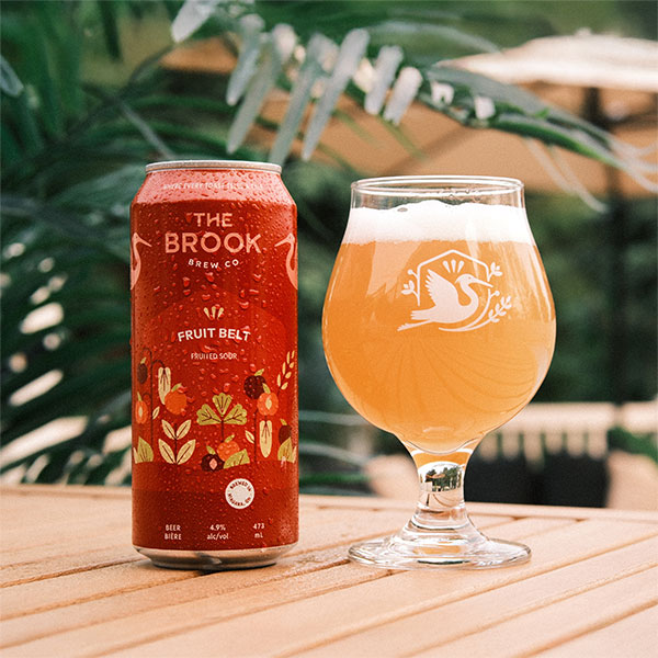 Fruit Belt Fruited Sour can and poured glass of beer from The Brook Brew Co. on a patio table with greenery in the background