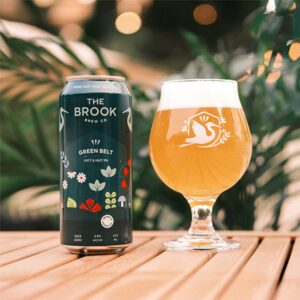 Green Belt Juicy & Hazy IPA can and poured glass of beer from The Brook Brew Co. on a wooden table with plants in the background