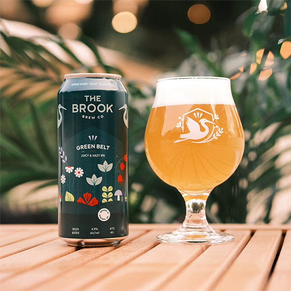 Green Belt Juicy & Hazy IPA can and poured glass of beer from The Brook Brew Co. on a wooden table with plants in the background