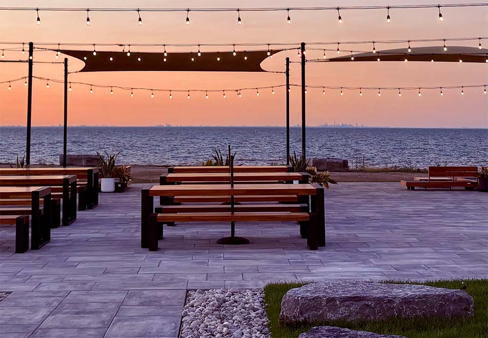 Outdoor beer garden patio with wooden picnic tables, umbrellas and string lights overlooking Lake Ontario at dusk, the Toronto skyline in the distance.