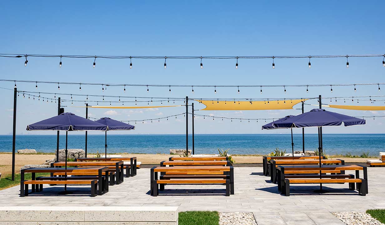 Outdoor beer garden patio with wooden picnic tables, umbrellas and string lights overlooking Lake Ontario.